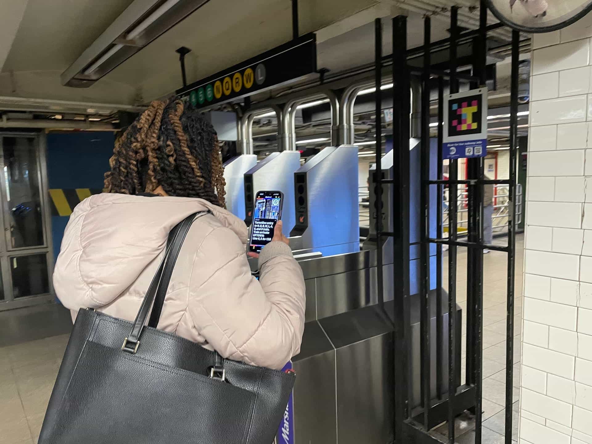 Pasajera en una estación de metro apuntando su teléfono hacia un código NaviLens ubicado junto a los torniquetes.